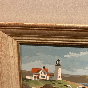 May include: A framed painting of a seascape with a lighthouse and a white house in the foreground. The sky is blue with white clouds. The painting is in a light wood frame.