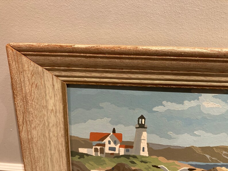May include: A framed painting of a seascape with a lighthouse and a white house in the foreground. The sky is blue with white clouds. The painting is in a light wood frame.