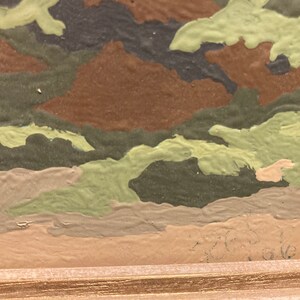 May include: Close-up of a framed painting with a camouflage pattern in shades of green, brown, and beige. The paint is textured and appears to be applied with a brush.