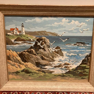 May include: A framed painting of a seascape with a lighthouse in the distance. The painting features a rocky coastline with crashing waves and seagulls flying overhead. The sky is a light blue with white clouds.