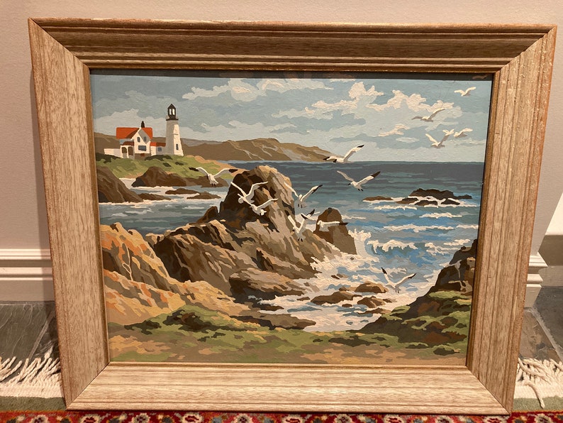 May include: A framed painting of a seascape with a lighthouse in the distance. The painting features a rocky coastline with crashing waves and seagulls flying overhead. The sky is a light blue with white clouds.