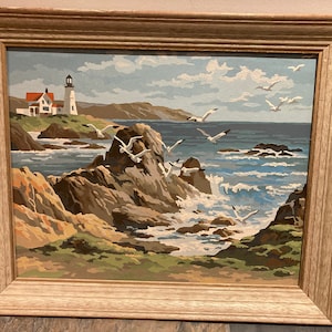 May include: A framed painting of a seascape with a lighthouse on a rocky shore. The painting features a blue sky with white clouds, a blue ocean with white waves, and brown rocks with green grass. There are white seagulls flying in the sky and over the water.