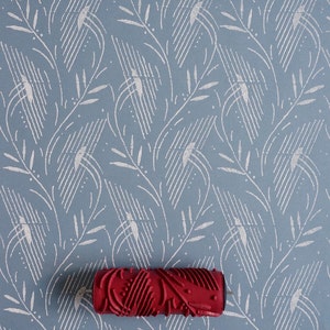 May include: A red patterned paint roller lies on a blue and white patterned surface. The surface features a repeating design of white lines and leaf-like shapes. The roller has a detailed, textured pattern. The image is a top-down view.