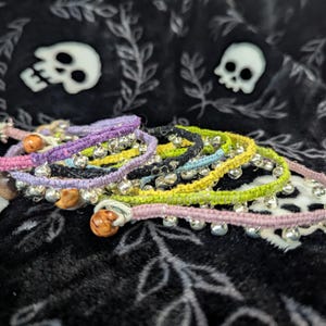 May include: A collection of colorful, woven anklets with small silver bells and wooden beads. The anklets are in shades of pink, purple, yellow, green, and black. The background is a black fabric with white skull and leaf patterns.