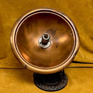 May include: A vintage copper-colored lamp shade with a circular design. The shade has a bowl shape with a central fixture. The base is a dark, ornate metal. The lamp is set against a yellow-gold background.