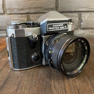 May include: A black and silver 35mm film camera with a silver top plate and a black lens. The camera has a black leatherette covering and a silver lens ring. The camera is labeled "HANIMEX PRAKTICA super TL" and the lens is labeled "AUTOMATIC 50mm 1:2.8" and "MADE IN JAPAN".