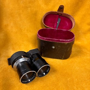 Puede incluir: Un juego de lentes de cámara negras vintage con un estuche de cuero a juego. Las lentes tienen marcas plateadas y blancas. El estuche es marrón oscuro con un interior de terciopelo rojo y un pequeño cierre.