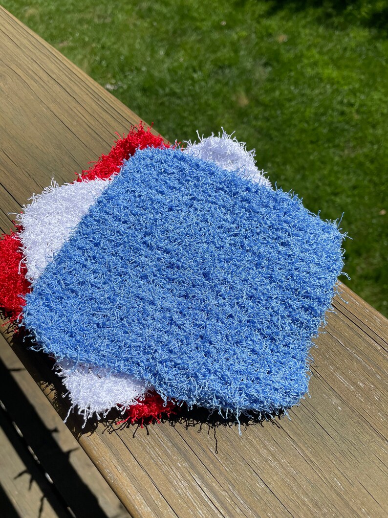 May include: Three crocheted dishcloths in blue, white, and red. The dishcloths are made with a fluffy, textured yarn.