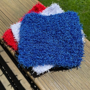May include: Three crocheted dishcloths in red, white, and blue. The cloths are made of a fluffy, textured yarn.