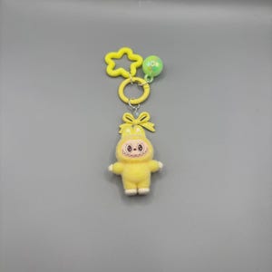 May include: A yellow plush keychain featuring a cartoon character with a bow and a smiling face. The keychain includes a star-shaped and a round charm, all in yellow and green. The character has white feet and a white belly. The keychain is attached to a yellow ring.