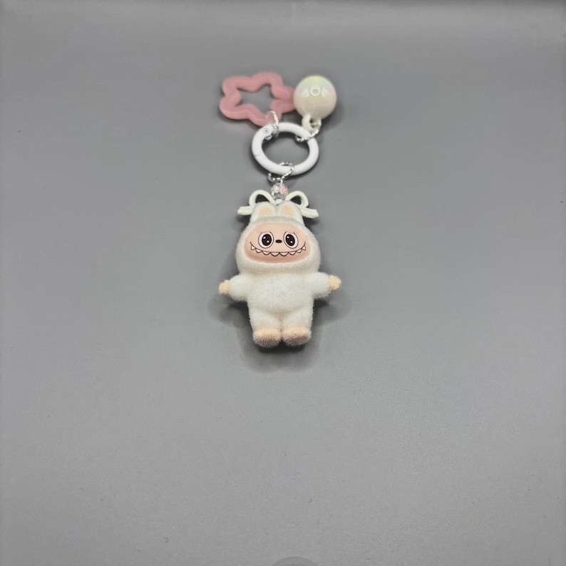 Puede incluir: Un llavero de felpa blanco con un personaje de dibujos animados sonriente con una cabeza redonda y brazos y piernas peque&ntilde;os. El llavero incluye un dije en forma de estrella rosa, un anillo blanco y una peque&ntilde;a bola blanca con el texto "40".