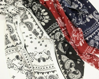 May include: Three hair scrunchies with long, flowing tails. The scrunchies are made of a white fabric with a black paisley pattern. The tails are tied in a knot at the top and have a frayed edge.