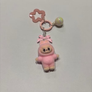 Può includere: Un portachiavi rosa con una figura di peluche con un volto sorridente e un fiocco. Il portachiavi include un ciondolo a forma di stella e una perla perlata. La figura indossa un completo rosa con cappuccio.