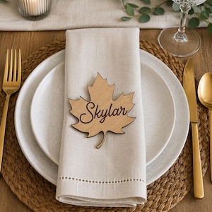 May include: A light-colored wooden place card shaped like a maple leaf with the name Skylar engraved in a dark cursive script. The leaf is positioned on a folded beige linen napkin resting on a white ceramic plate atop a woven round placemat.