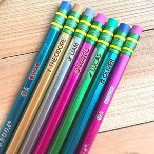 May include: A set of six metallic pencils with erasers, each featuring a unique name and symbol printed on the barrel. The pencils are colored in shades of blue, gold, silver, magenta, green, and purple, with green and yellow striped ferrules.