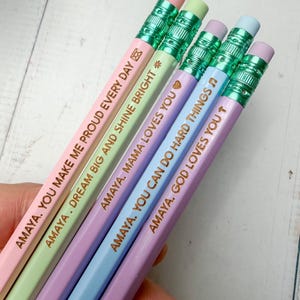 May include: Five pastel-colored pencils with metallic green ferrules and erasers. Each pencil features gold-stamped text: AMAYA. YOU MAKE ME PROUD EVERY DAY, AMAYA. DREAM BIG AND SHINE BRIGHT, AMAYA. MAMA LOVES YOU, AMAYA. YOU CAN DO HARD THINGS, and AMAYA. GOD LOVES YOU.