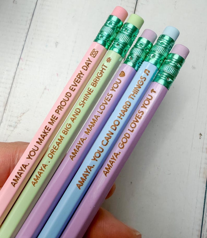 May include: Five pastel-colored pencils with metallic green ferrules and erasers. Each pencil features gold-stamped text: AMAYA. YOU MAKE ME PROUD EVERY DAY, AMAYA. DREAM BIG AND SHINE BRIGHT, AMAYA. MAMA LOVES YOU, AMAYA. YOU CAN DO HARD THINGS, and AMAYA. GOD LOVES YOU.