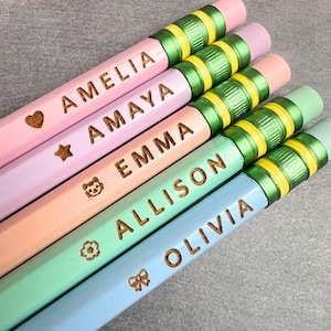 May include: Five pastel-colored pencils with metallic green and yellow ferrules and pink erasers. Each pencil is engraved with a name and a small icon: AMELIA with a heart, AMAYA with a star, EMMA with a cat, ALLISON with a flower, and OLIVIA with a bow.