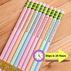 May include: A collection of pastel-colored pencils, each inscribed with "KYLIE DONALDSON" and adorned with a star and heart. The pencils have green and yellow striped erasers. The pencils are bundled together with a gold glitter ribbon.