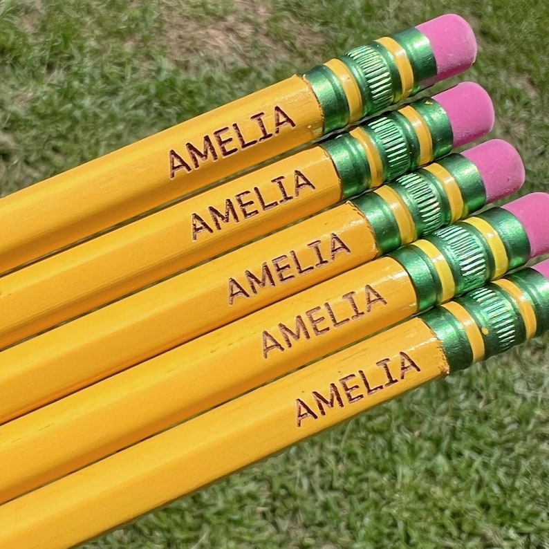 May include: Five yellow wooden pencils arranged in a parallel stack. Each pencil features the name AMELIA engraved in black capital letters. The pencils have a metallic green ferrule and a pink eraser at the top end of each writing instrument.