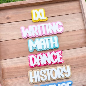 May include: A set of layered acrylic signs with white raised lettering on colorful backgrounds. The signs feature the words IXL in yellow, WRITING in pink, MATH in light blue, DANCE in a pink star pattern, and HISTORY in a natural wood finish.