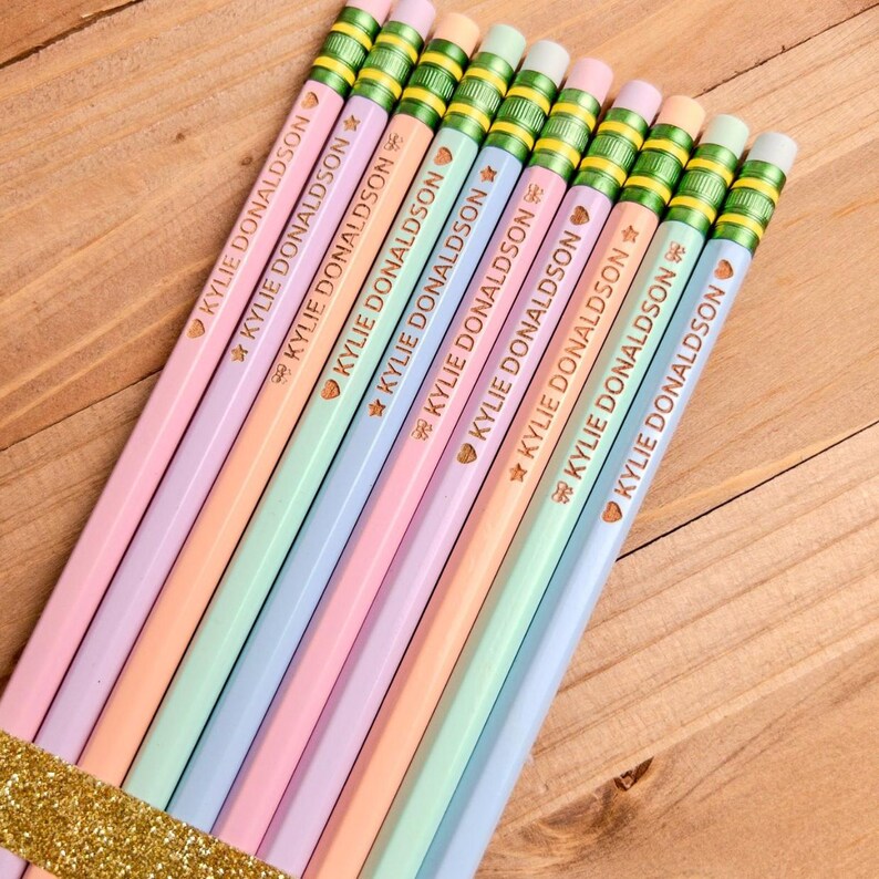 May include: A set of nine pastel-colored pencils arranged in a row, each featuring a metallic green ferrule and a pink eraser. The pencils are imprinted with the name KYLIE DONALDSON in gold lettering, accompanied by a small gold heart icon.