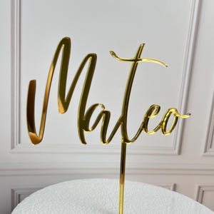 May include: A gold-colored cake topper with the name "Mateo" in a cursive font. The topper is made of a reflective material and is mounted on a thin, gold-colored stem. The background is a blurred white.