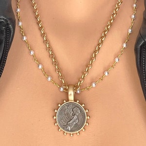 May include: A gold chain necklace with a pearl chain layered on top. The necklace features a pendant with a circular design and a depiction of an angel with wings.