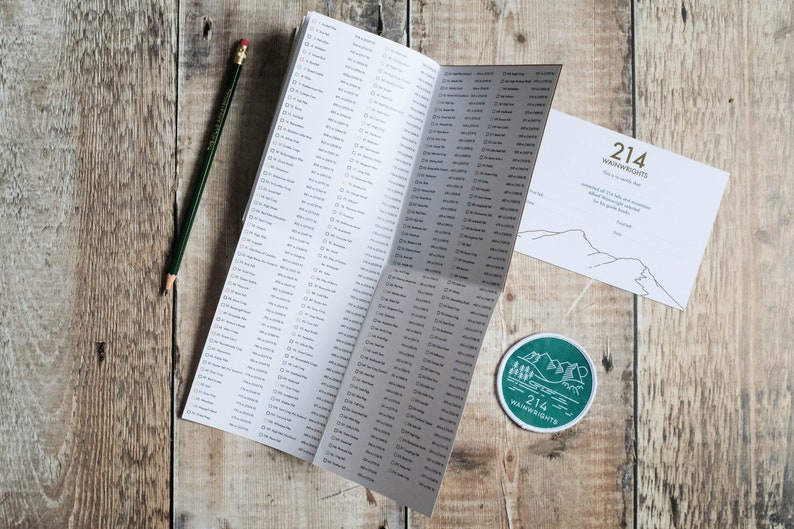 K&ouml;nnte beinhalten: Eine Checkliste der 214 Wainwright-Fells im Lake District, England. Die Checkliste ist auf einer Seite mit einer Liste der Fells ge&ouml;ffnet. Ein runder Aufkleber in Gr&uuml;n und Wei&szlig; mit dem Text "214 Wainwrights" befindet sich auf einer Holzoberfl&auml;che. Eine wei&szlig;e Karte mit dem Text "214 Wainwrights" und einer Strichzeichnung von Bergen befindet sich ebenfalls auf der Oberfl&auml;che.