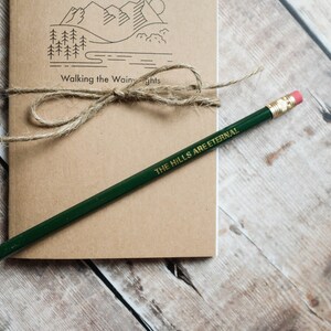 K&ouml;nnte beinhalten: Ein braunes Notizbuch mit einer Zeichnung von Bergen und B&auml;umen auf dem Einband. Das Notizbuch ist mit Schnur gebunden und tr&auml;gt den Text "Walking the Wainwright" auf dem Einband. Ein gr&uuml;ner Bleistift mit dem Text "THE HILLS ARE ETERNAL" liegt auf dem Notizbuch.