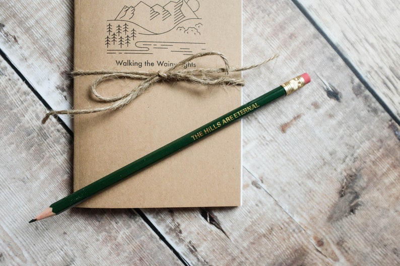 K&ouml;nnte beinhalten: Ein braunes Notizbuch mit einer Zeichnung von Bergen und B&auml;umen auf dem Einband. Das Notizbuch ist mit Schnur gebunden und tr&auml;gt den Text "Walking the Wainwright" auf dem Einband. Ein gr&uuml;ner Bleistift mit dem Text "THE HILLS ARE ETERNAL" liegt auf dem Notizbuch.