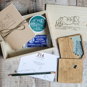 K&ouml;nnte beinhalten: Eine Holzkiste gef&uuml;llt mit einem braunen Notizbuch, einem gr&uuml;nen Patch mit dem Text "214 Wainwrights", einem blau-wei&szlig;en Schokoriegel und zwei Holz-Untersetzern mit einem topografischen Kartenmotiv. Die Kiste steht auf einer Holzoberfl&auml;che.