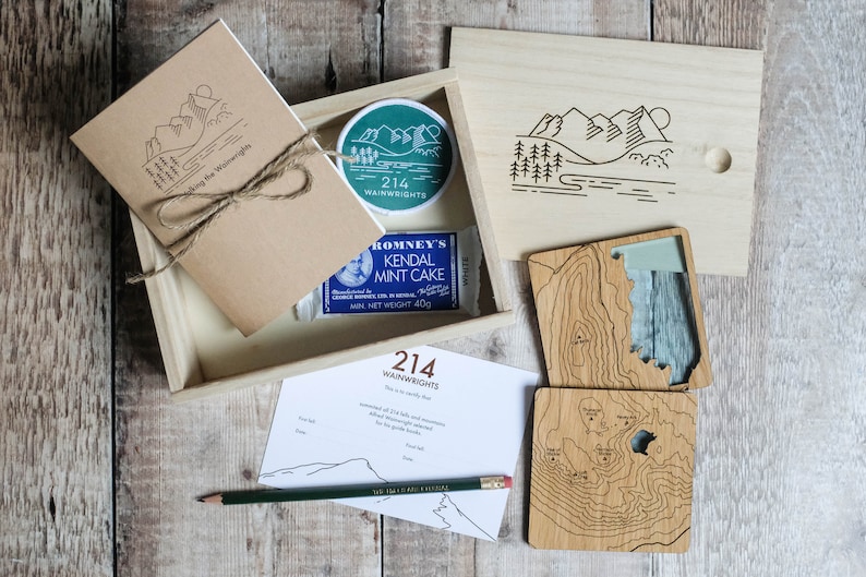 K&ouml;nnte beinhalten: Eine Holzkiste gef&uuml;llt mit einem braunen Notizbuch, einem gr&uuml;nen Patch mit dem Text "214 Wainwrights", einem blau-wei&szlig;en Schokoriegel und zwei Holz-Untersetzern mit einem topografischen Kartenmotiv. Die Kiste steht auf einer Holzoberfl&auml;che.