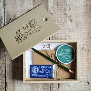 K&ouml;nnte beinhalten: Eine Holzkiste mit einem Scharnierdeckel, der Deckel ist ge&ouml;ffnet und zeigt eine Bergszene mit Sonne und B&auml;umen. In der Kiste befinden sich ein gr&uuml;ner Aufn&auml;her mit dem Text "214 WAINWRIGHTS", ein brauner Papierumschlag, ein gr&uuml;ner Bleistift, ein wei&szlig;er Radiergummi und eine blaue und wei&szlig;e Packung Romney's Kendal Mint Cake.