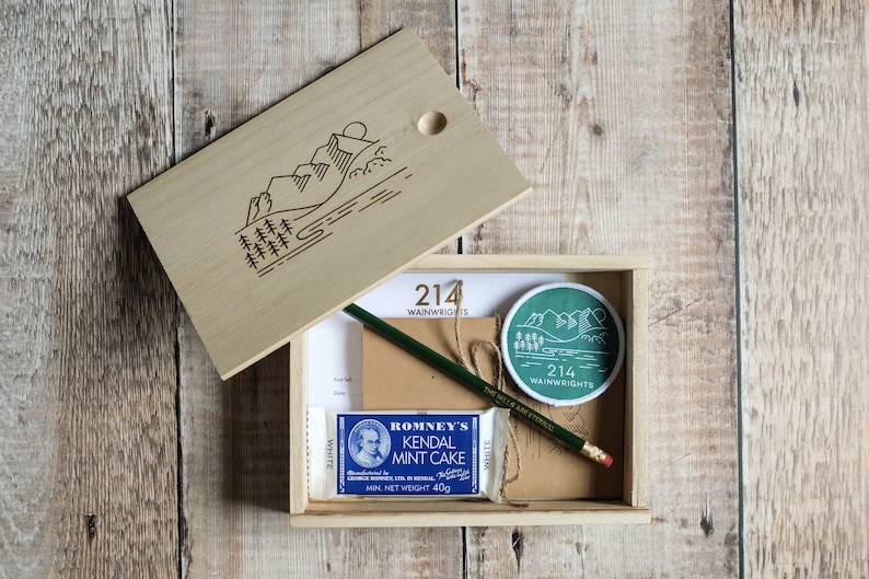 K&ouml;nnte beinhalten: Eine Holzkiste mit einem Scharnierdeckel, der Deckel ist ge&ouml;ffnet und zeigt eine Bergszene mit Sonne und B&auml;umen. In der Kiste befinden sich ein gr&uuml;ner Aufn&auml;her mit dem Text "214 WAINWRIGHTS", ein brauner Papierumschlag, ein gr&uuml;ner Bleistift, ein wei&szlig;er Radiergummi und eine blaue und wei&szlig;e Packung Romney's Kendal Mint Cake.