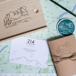 K&ouml;nnte beinhalten: Eine Holzkiste mit einem eingravierten Bergketten-Design auf dem Deckel. Die Kiste enth&auml;lt ein Zertifikat mit dem Text "214 Wainwrights This is to certify that summited all 214 fells and mountains Alfred Wainwright selected for his guide books." Die Kiste enth&auml;lt au&szlig;erdem ein kleines braunes Notizbuch mit dem Text "Walking the Wainwrights" auf dem Umschlag.