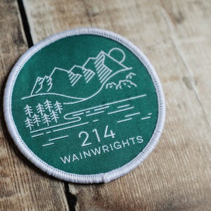 K&ouml;nnte beinhalten: Ein gr&uuml;ner runder Patch mit wei&szlig;en N&auml;hten am Rand. Der Patch zeigt eine wei&szlig;e Umrandung einer Bergkette, eines Sees und von B&auml;umen. Der Text "214 Wainwrights" ist unten auf dem Patch in Wei&szlig; gedruckt.