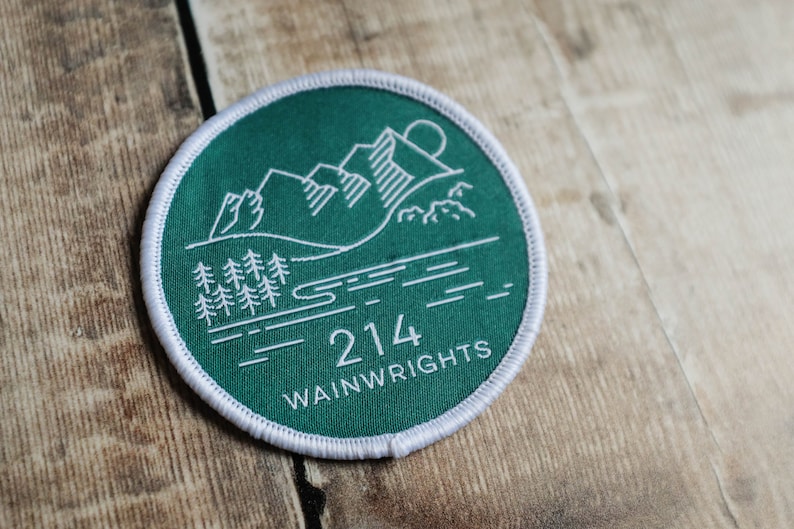K&ouml;nnte beinhalten: Ein gr&uuml;ner runder Patch mit wei&szlig;en N&auml;hten am Rand. Der Patch zeigt eine wei&szlig;e Umrandung einer Bergkette, eines Sees und von B&auml;umen. Der Text "214 Wainwrights" ist unten auf dem Patch in Wei&szlig; gedruckt.