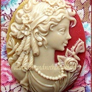 May include: A cameo brooch featuring a woman with a floral crown and a pearl necklace. The cameo is made of white and red resin and has a detailed design.