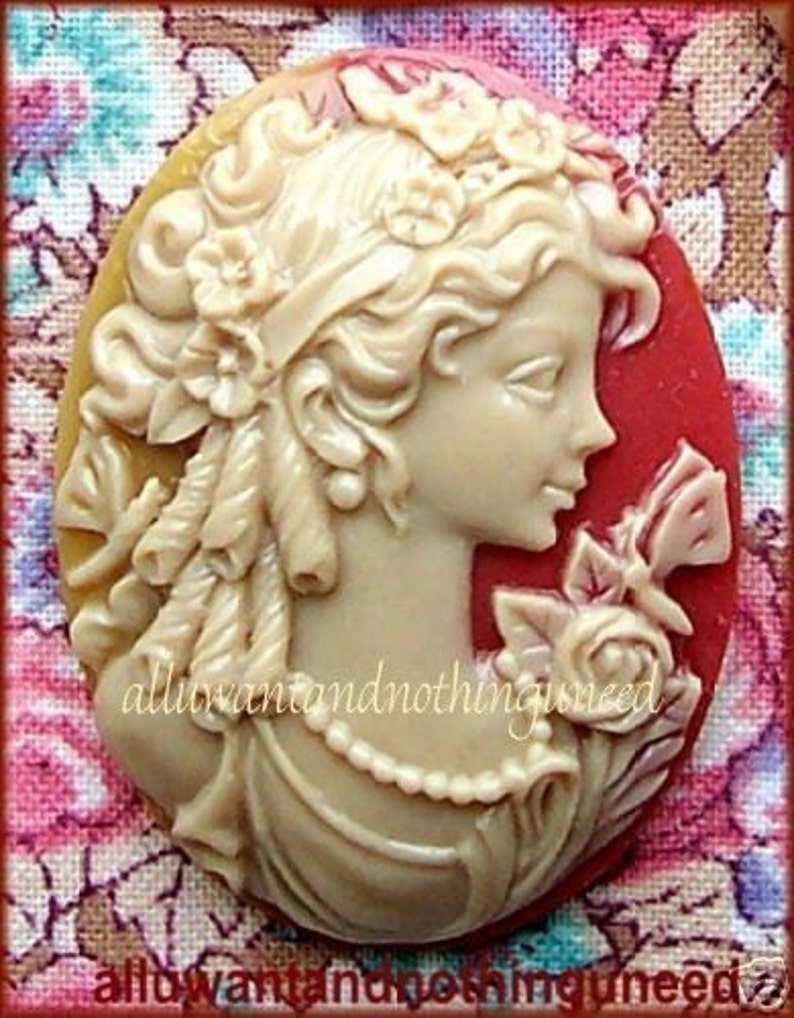 May include: A cameo brooch featuring a woman with a floral crown and a pearl necklace. The cameo is made of white and red resin and has a detailed design.