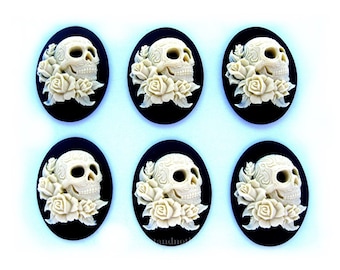 May include: Six white cameo skull and rose cabochons with black backgrounds. The skulls are detailed with intricate designs and are surrounded by white roses.