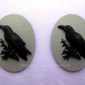 May include: Two gray oval-shaped cameo pendants with black raven silhouettes perched on branches. The ravens are facing to the right.