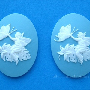 May include: Two blue and white ceramic cameo pendants featuring a fairy with butterfly wings holding a butterfly. The fairy is sitting on a leaf with a flower.
