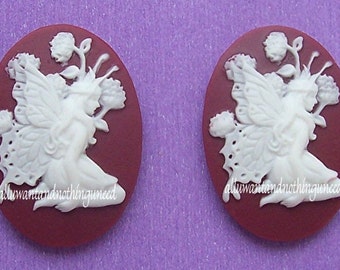 May include: Two oval-shaped cameo pendants. Each features a white fairy design with detailed wings and floral accents, set against a burgundy background. The fairy is in profile, holding flowers. The text "alluwantandnothinguneed" is at the bottom.