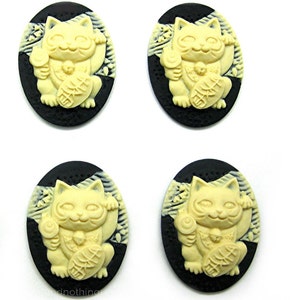 May include: Four oval-shaped cameo pendants featuring a cream-colored, smiling Maneki-neko cat design on a black background. The cat is holding a gold coin and waving its paw. The pendants are arranged on a white surface.