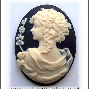 May include: A white cameo featuring a woman with curly hair, wearing a draped garment, and a floral design on the side. The cameo is set against a black background.