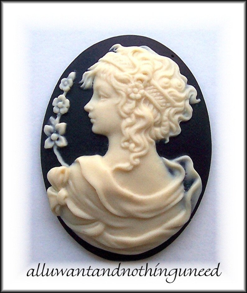 May include: A white cameo featuring a woman with curly hair, wearing a draped garment, and a floral design on the side. The cameo is set against a black background.