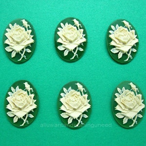 May include: Six white cameo-style rose flower embellishments with green leaves on a green background.