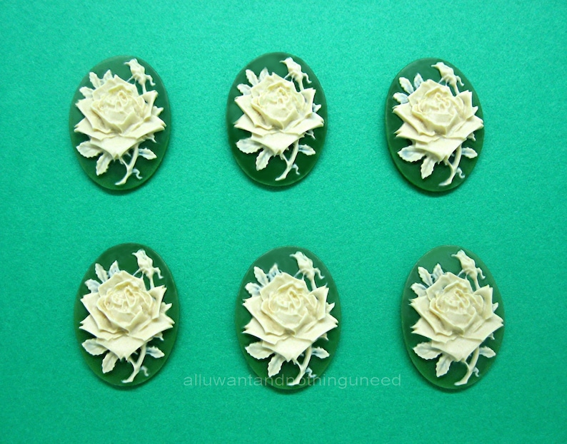 May include: Six white cameo-style rose flower embellishments with green leaves on a green background.