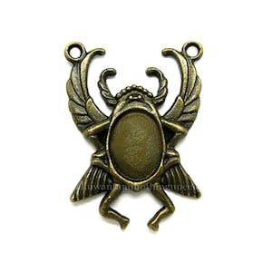 May include: A bronze-colored metal pendant shaped like a scarab beetle with wings outstretched. The pendant has a round, empty space in the center for adding a cameo or other embellishment.